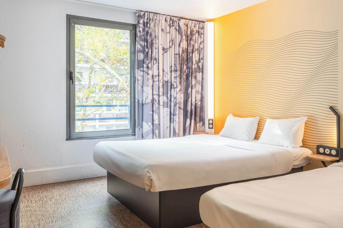 A hotel room with a large bed, a bedside table with a lamp, a window with curtains, and a view of trees outside.