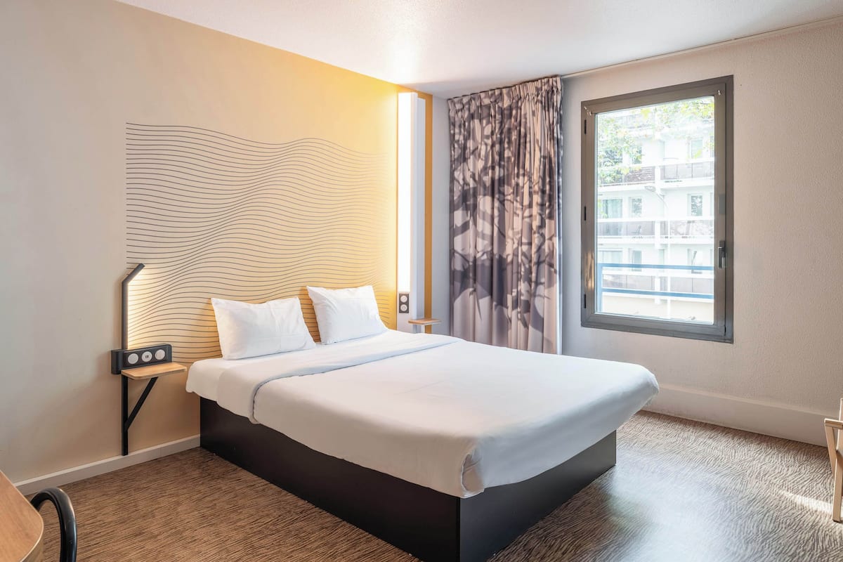 A hotel room with a bed, a nightstand, a lamp, a window with curtains, and a view of buildings outside.