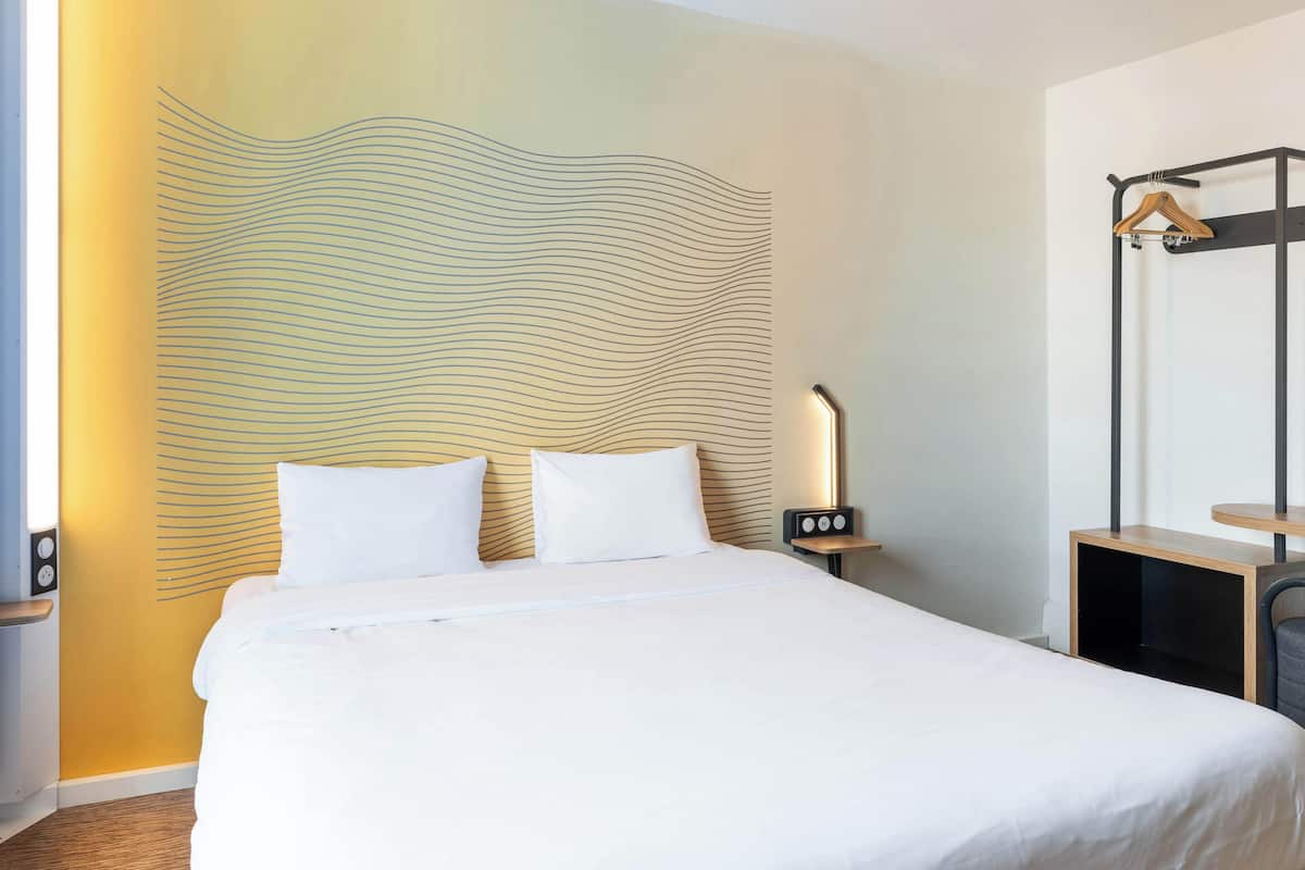 A neatly made bed with white linens, a bedside table with a lamp, and a wall-mounted shelf.