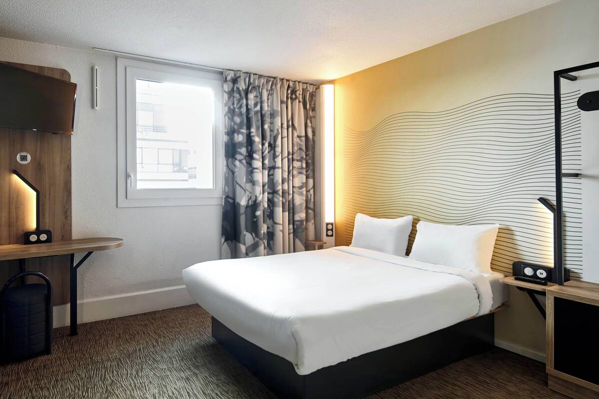 Habitación de hotel con una cama grande, un escritorio, una silla, un ventanal con cortinas, una lámpara de pared y una obra de arte en la pared.