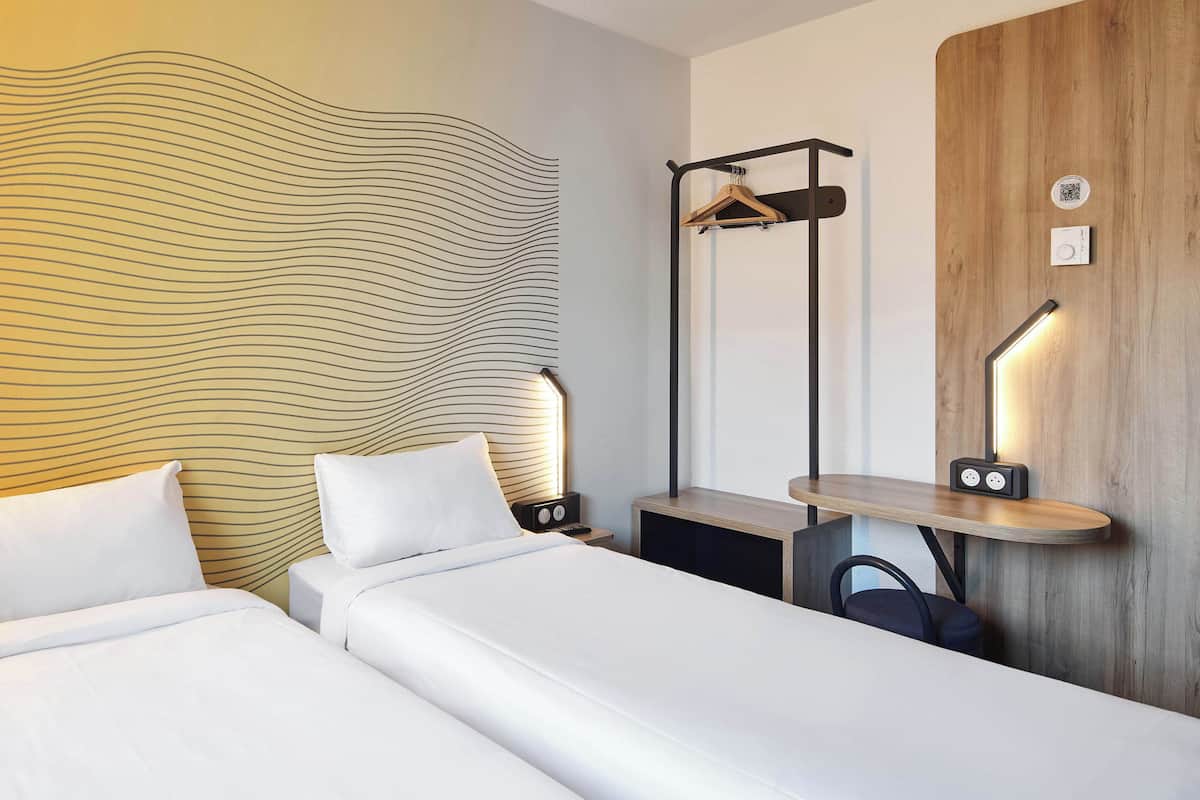 Habitación de hotel con dos camas, cabecera de madera, mesita de noche con lámpara, perchero y una pequeña estantería con un radio.