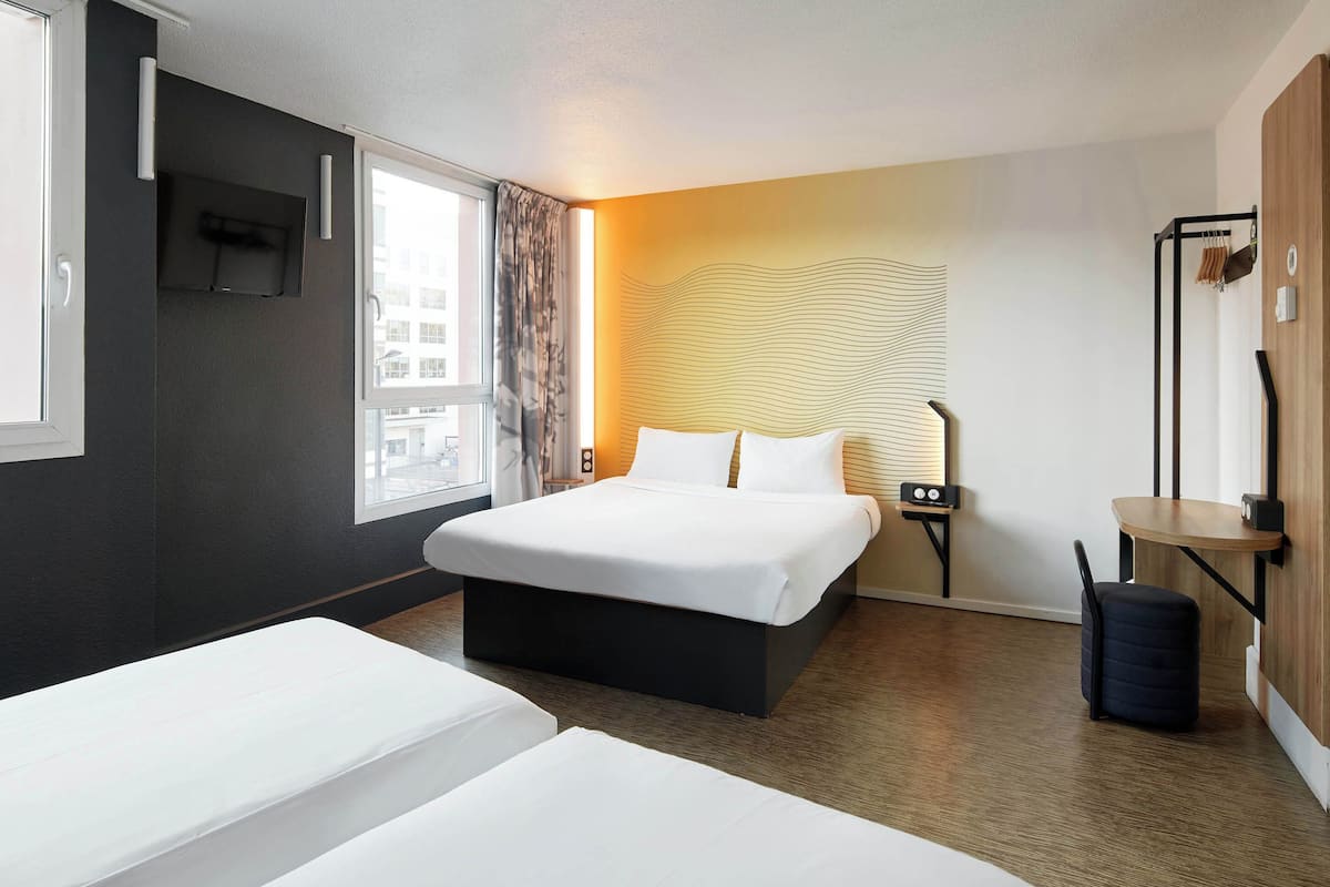 Habitación de hotel con una cama grande, una cama más pequeña, una mesita de noche, una lámpara, una maleta y vistas a la ciudad por la ventana.