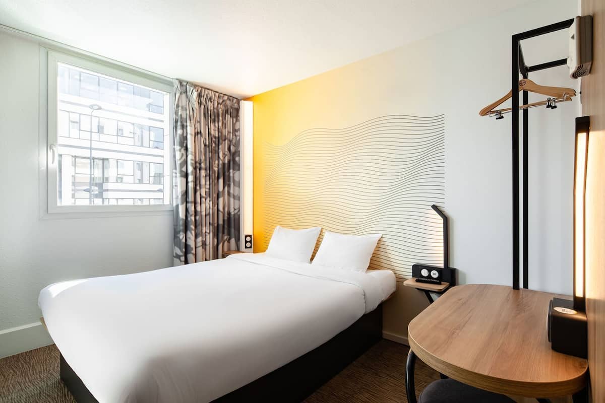 Habitación de hotel con cama, mesita de noche con lámpara, mesita auxiliar de madera y ventana con cortinas.