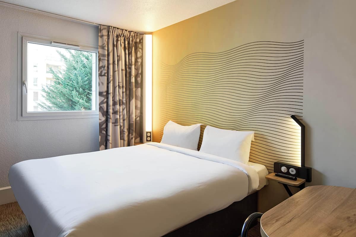 Habitación de hotel con una cama grande, una mesita de noche con lámpara, una pequeña mesa y vistas a árboles a través de la ventana.