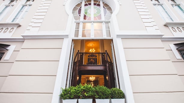 Eine Gebäudefassade mit einem großen Bogenfenster, einer Treppe zu einer oberen Ebene und einem kleinen Garten davor.