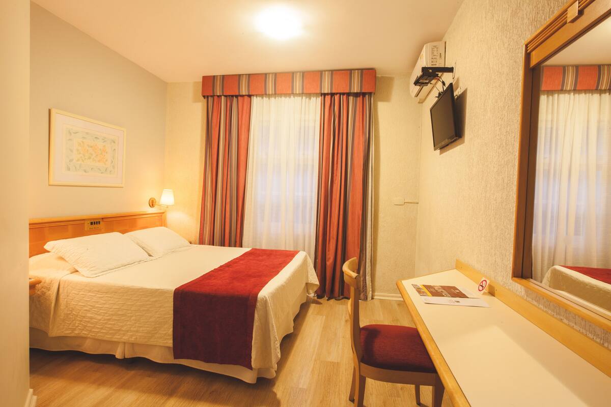 A hotel room with a bed, a desk, a chair, a television, and a window with curtains.