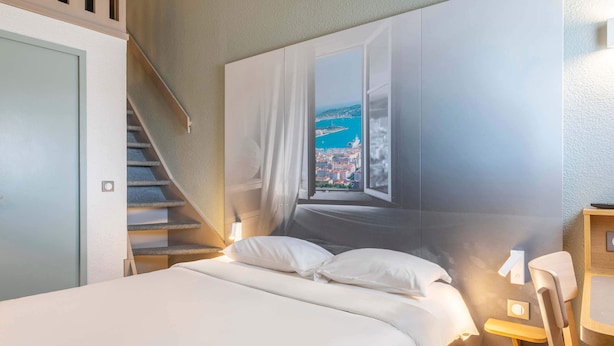 Une chambre à coucher comprenant un lit, une chaise, un bureau, une fenêtre donnant sur une ville côtière et un escalier.