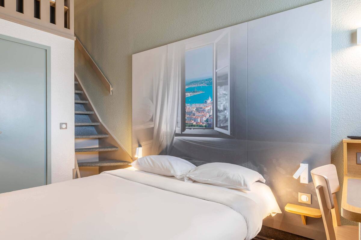 A bedroom with a bed, a chair, a desk, a window with a view of a coastal city, and a staircase.