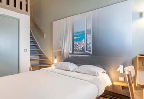 Une chambre à coucher comprenant un lit, une chaise, un bureau, une fenêtre donnant sur une ville côtière et un escalier.