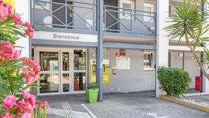 L’entrée d’un hôtel, avec une pancarte « Bienvenue », une place de stationnement accessible aux personnes à mobilité réduite et un bac à fleurs vert.