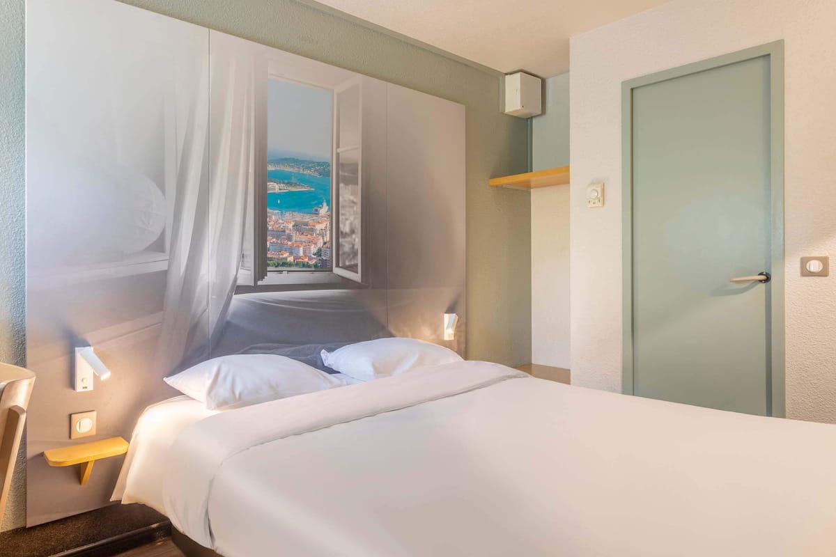 A hotel room with a bed, a nightstand, a wall-mounted light, a door, and a window with a view of a cityscape.