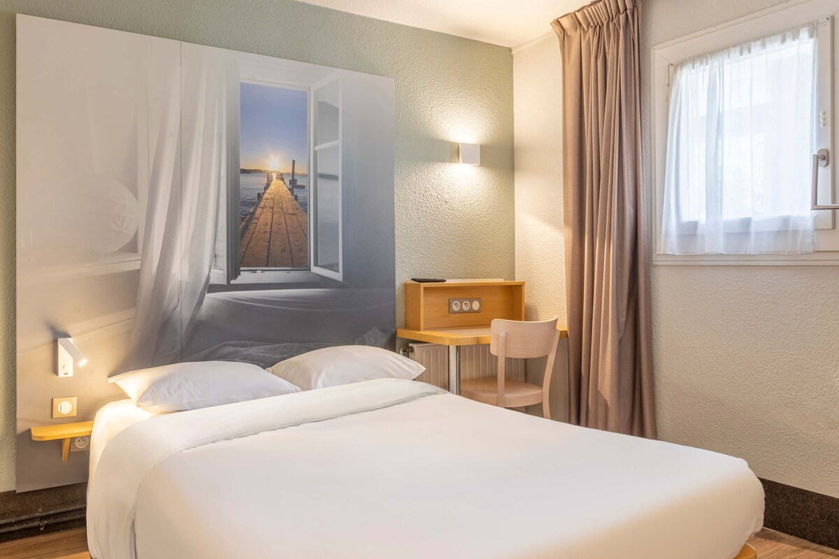 A hotel room with a large bed, a desk, a chair, and a wall-mounted TV.