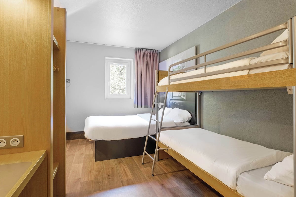 Una habitación compacta con dos literas, una puerta de madera, una ventana con cortinas y una mesita de noche pequeña.