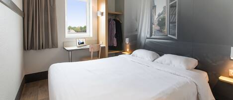 A hotel room with a large bed, a desk with a computer, a chair, a wardrobe, and a window with curtains.