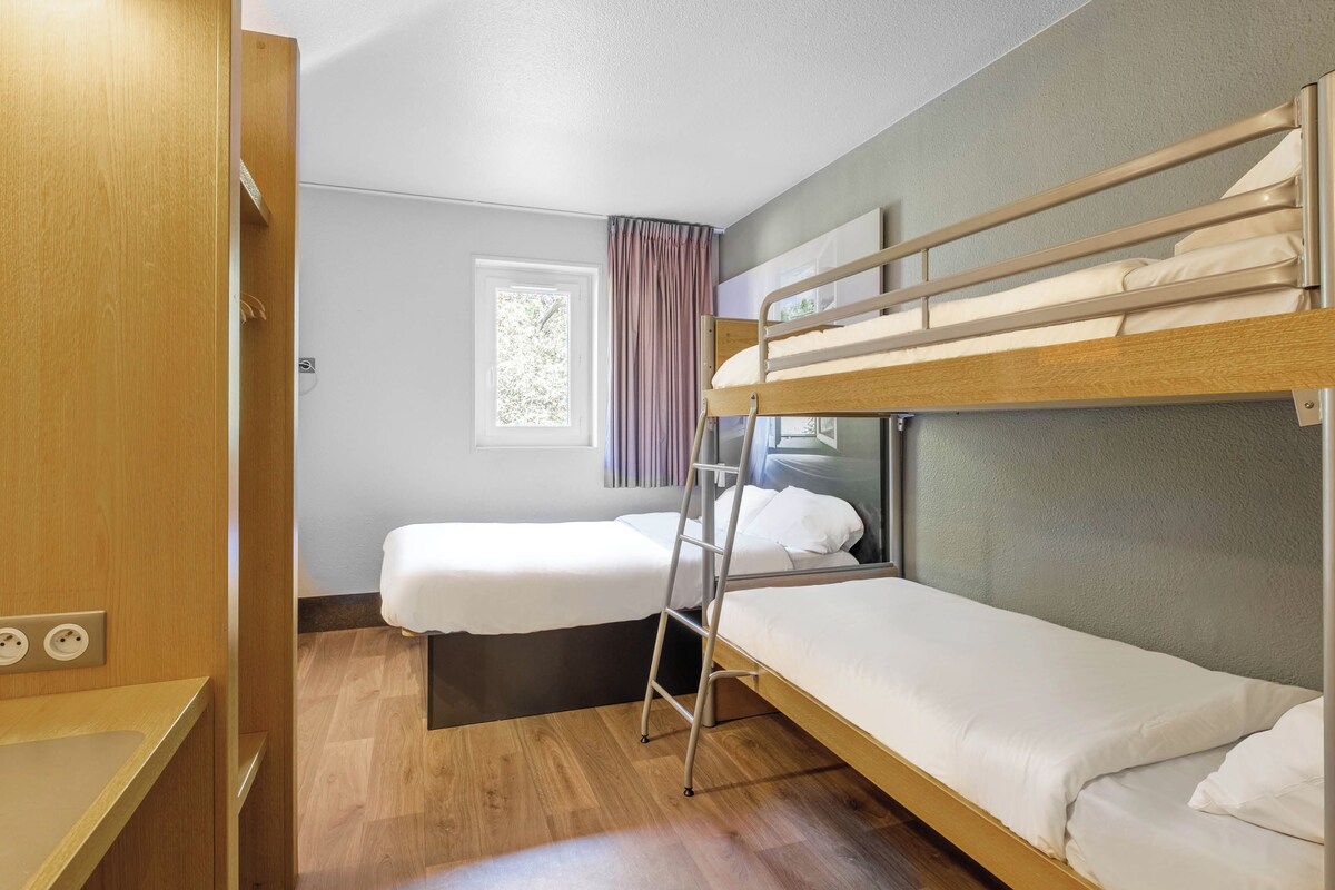 Una habitación compacta con dos literas, una puerta de madera, una ventana con cortinas y una mesita de noche pequeña.