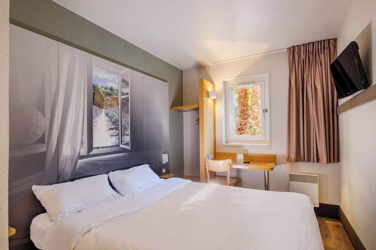 Habitación de hotel con cama, escritorio, silla, televisión y ventana con cortinas.