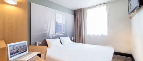 Une chambre d’hôtel avec un lit, un bureau avec un ordinateur portable, une télévision et une fenêtre avec des rideaux.
