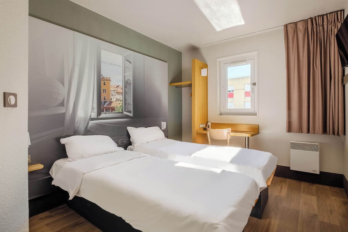 Habitación de hotel con dos camas, una ventana con cortinas, un escritorio con una silla y un aire acondicionado instalado en la pared.