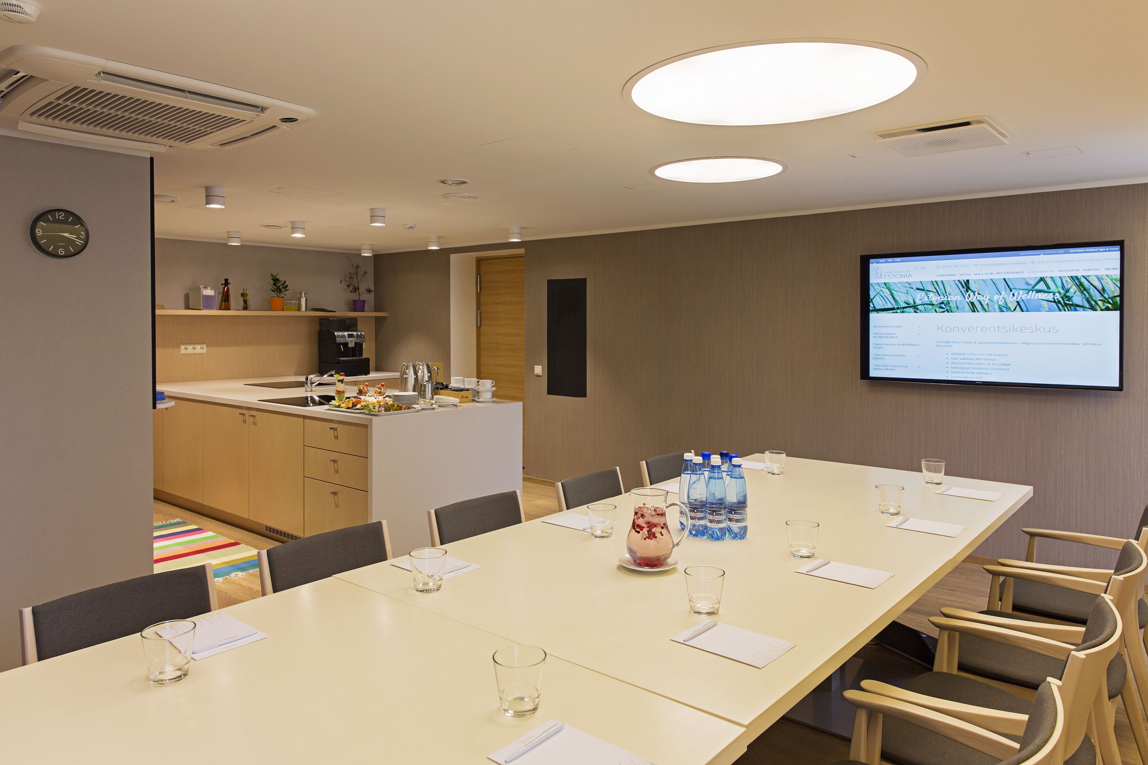 A conference room with a long table, chairs, a projector screen, and a kitchen area in the background.