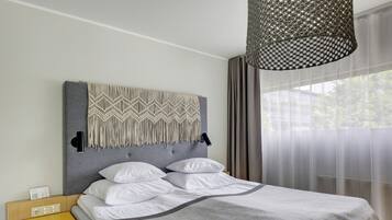 Una cama impecablemente hecha con colcha gris y almohadas blancas, cabecera gris con diseño de macramé, mesita de noche amarilla con toma de corriente y un ventanal con cortinas traslúcidas.