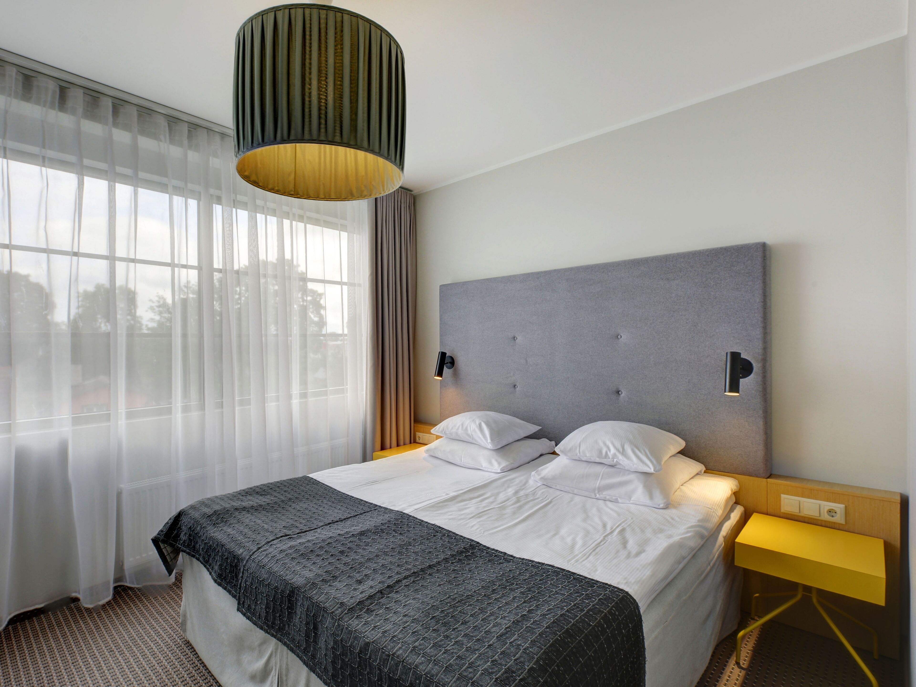 Una habitación de hotel con una cama, una mesita de noche, una lámpara, una ventana con cortinas y un cabecero.