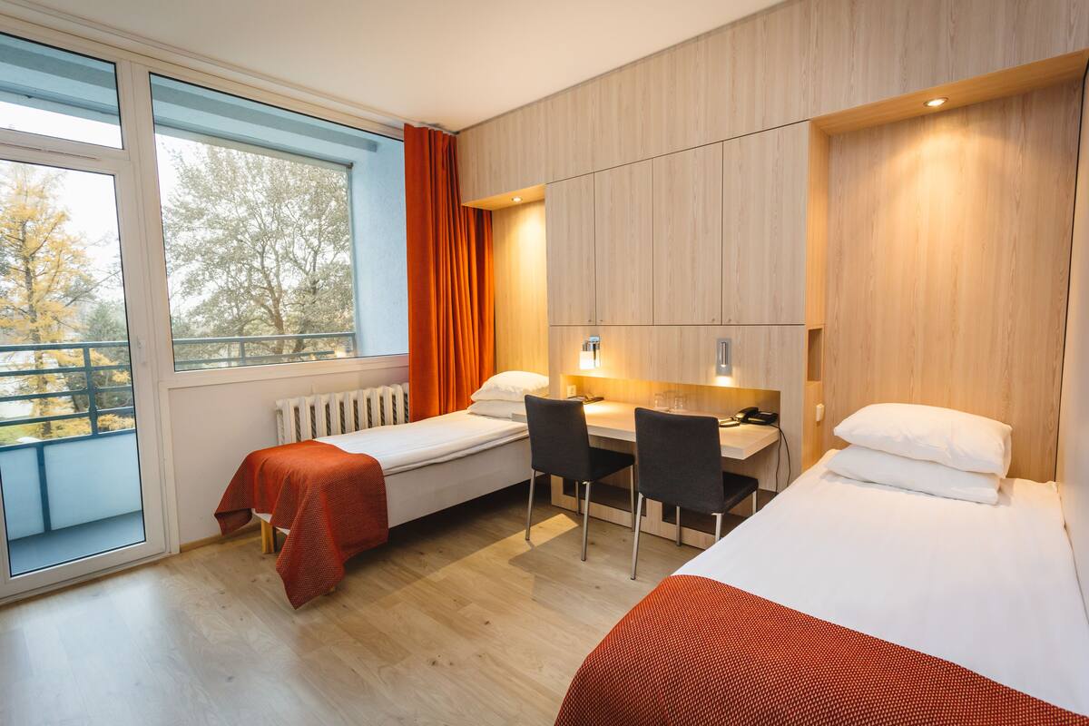 A modern hotel room with two single beds, a desk with chairs, a radiator, and a large window with curtains.