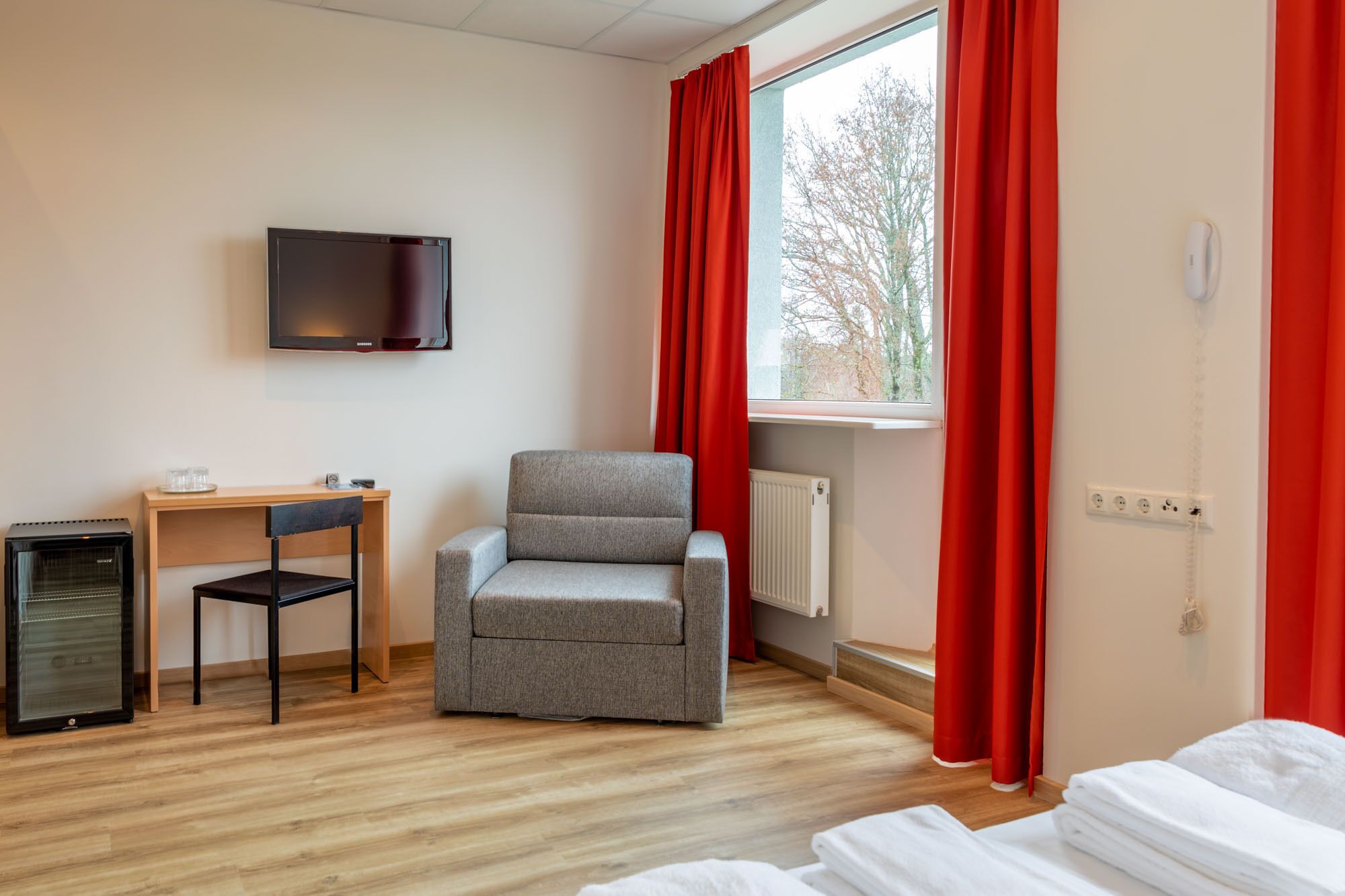 Ett hotellrum med en grå fåtölj, ett litet skrivbord med en stol, en tv som är monterad på väggen och ett fönster med röda gardiner.