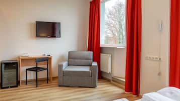 Ett hotellrum med en grå fåtölj, ett litet skrivbord med en stol, en tv som är monterad på väggen och ett fönster med röda gardiner.