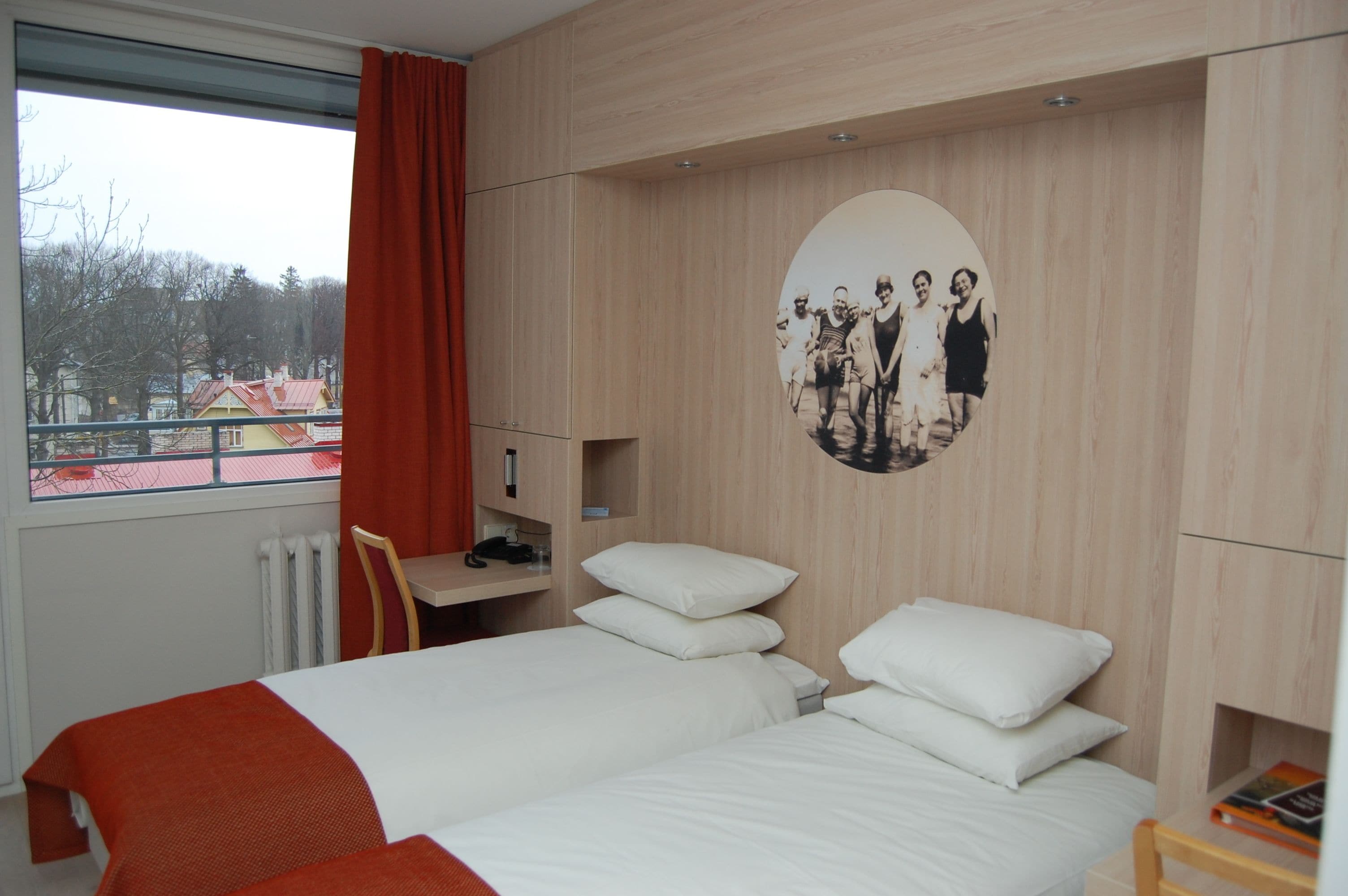 Een hotelkamer met twee bedden, een bureau, een stoel en een raam met uitzicht op gebouwen en bomen.