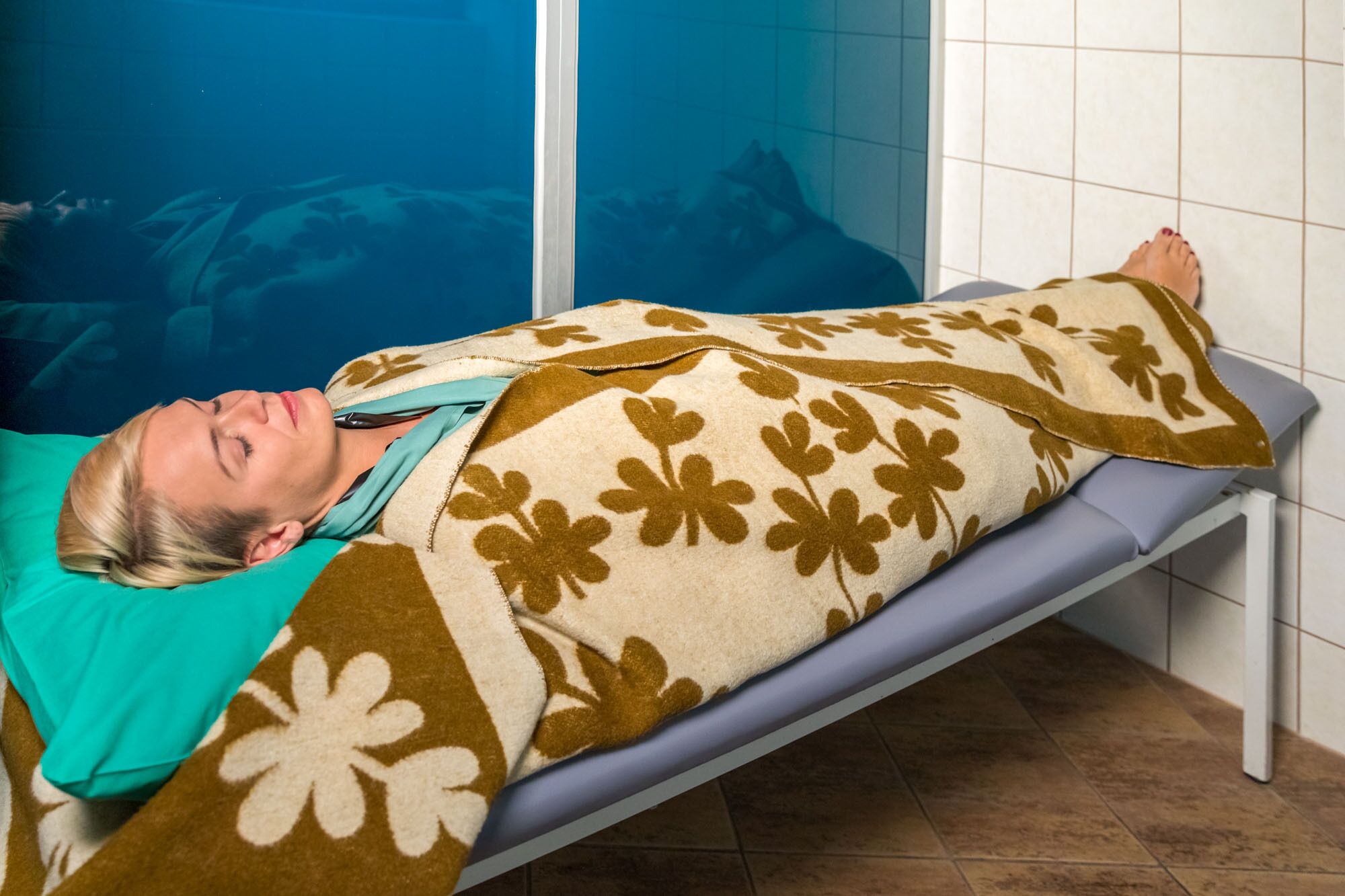 Una persona acostada en una cama en una habitación con pared azul y suelo de baldosas.