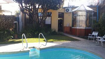 A swimming pool with steps leading into it, surrounded by a garden with various plants and a tree. There is a building with a yellow facade and a covered area with chairs.