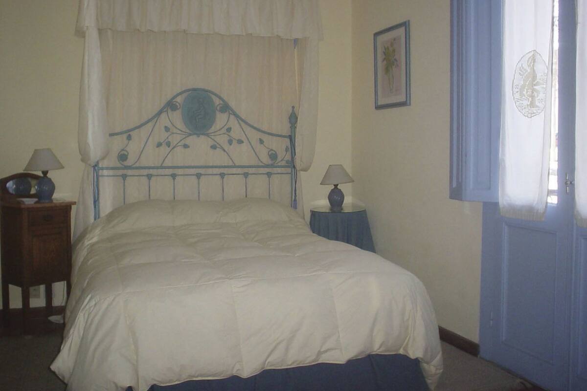 A bedroom with a bed, two bedside tables with lamps, a window with curtains, and a framed picture on the wall.