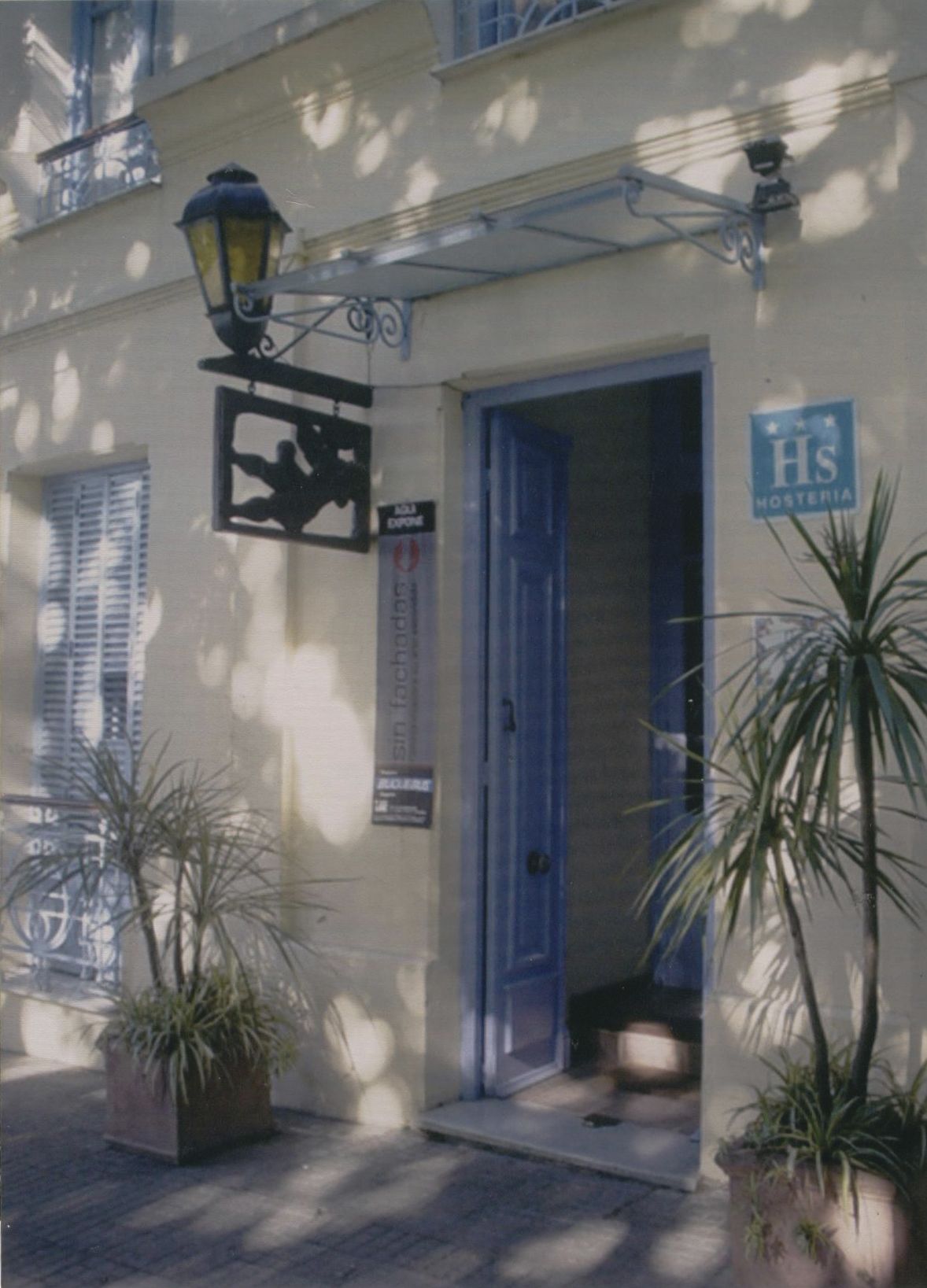 A building with a blue door, a sign reading "Hs Hosteria," and potted plants in front.