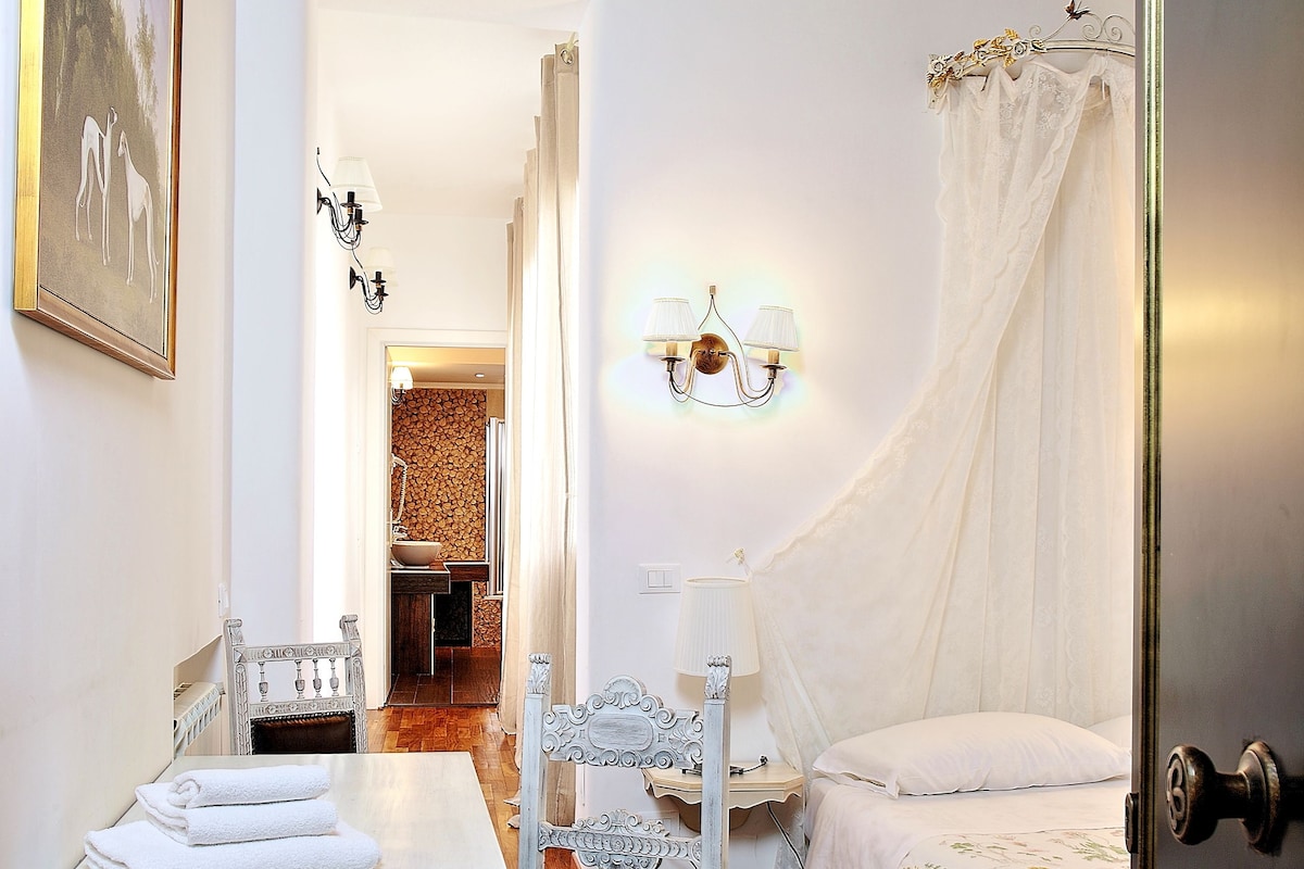 A bedroom with a bed, a bedside table, a chair, a table with towels, a wall-mounted lamp, and a door.