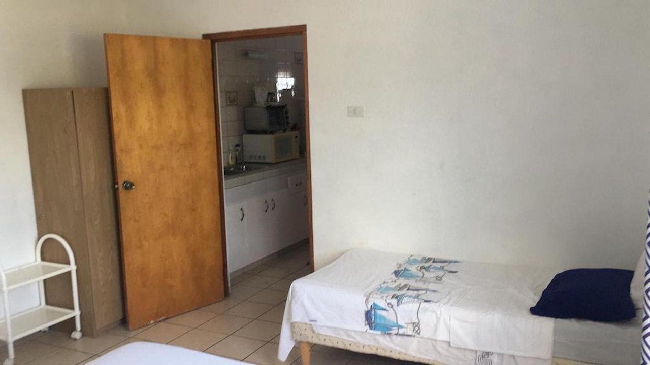 A bedroom with a bed, a wooden wardrobe, and a kitchenette in the background.