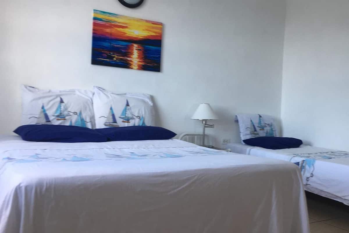 Une chambre d’hôtel avec deux lits, chacun agrémenté d’une couvre-lit orné d’un motif de voilier, une table de chevet, une lampe et un tableau représentant un coucher de soleil accroché au mur.
