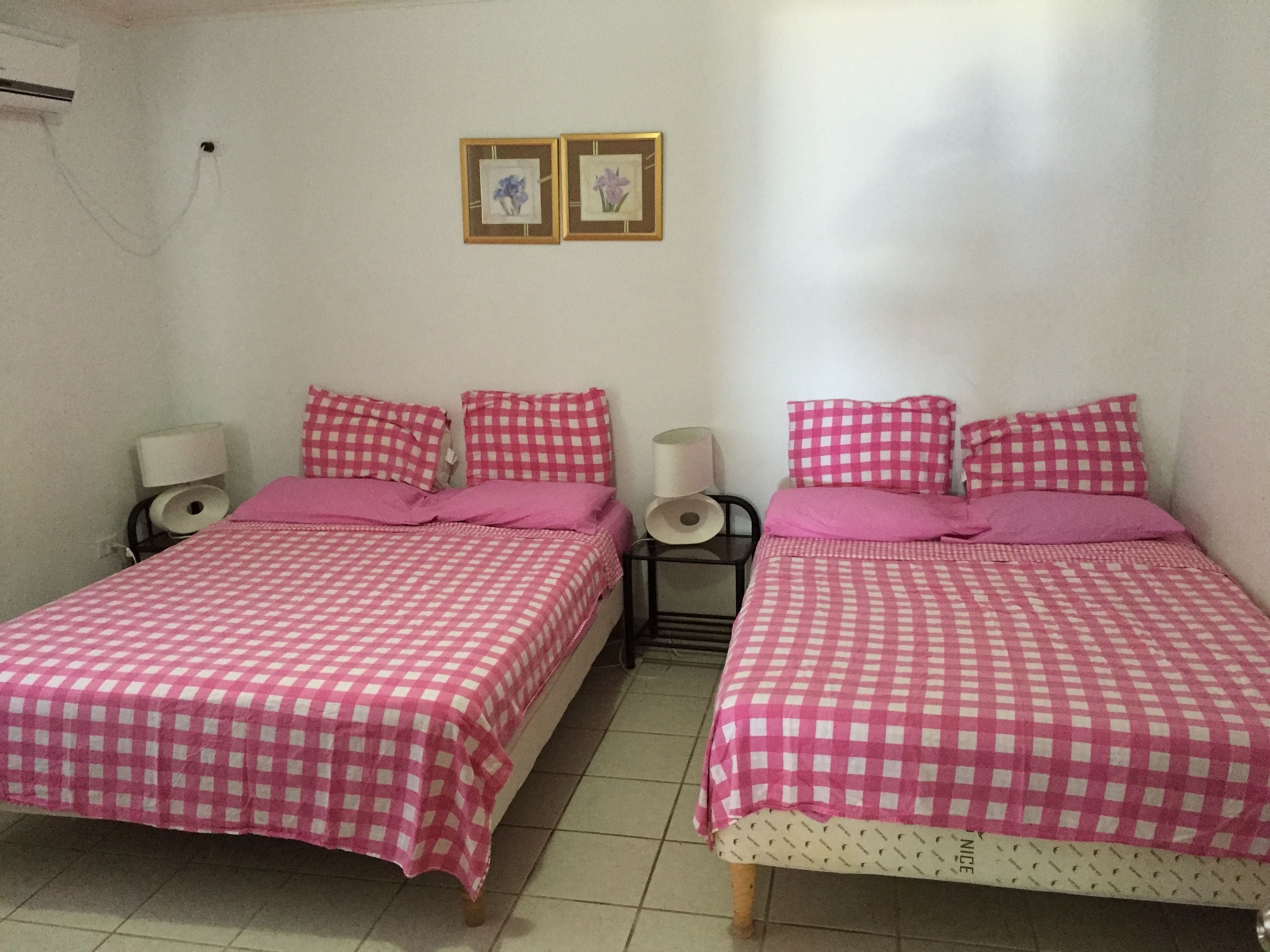 A room with two beds, each with a pink checkered bedspread and matching pillows. There are two framed pictures on the wall above the beds.
