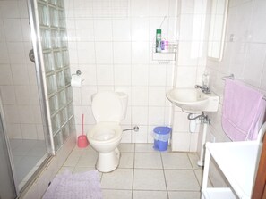 A bathroom with a toilet, sink, and shower. There is a shelf with toiletries and a pink towel hanging on the wall. - My Dream Apartments (Lagun)
