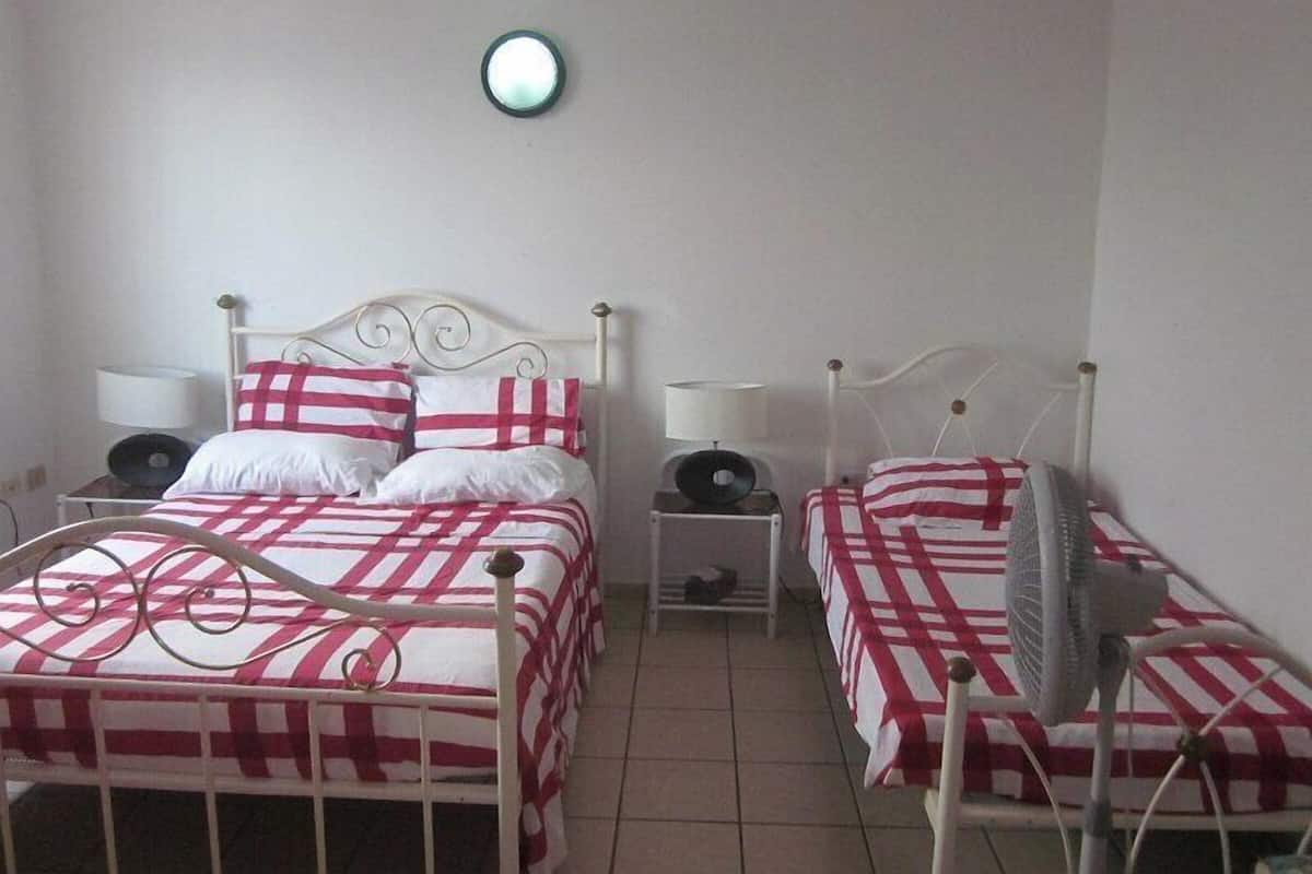 Une chambre avec deux lits, chacun doté d’une structure en métal blanc et d’une literie à carreaux rouges et blancs. On y trouve des tables de chevet avec des lampes, ainsi qu’un luminaire fixé au mur.