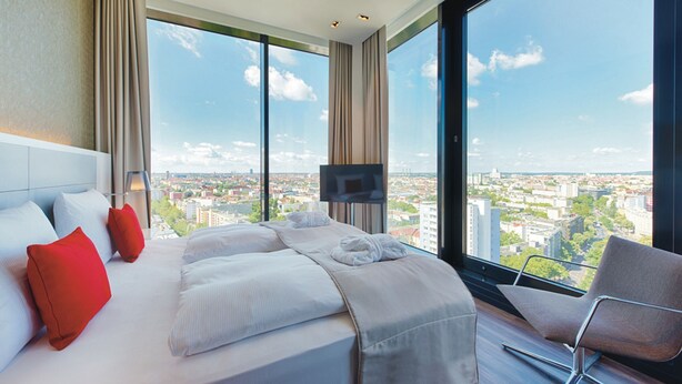 Una habitación de hotel moderna con una cama grande, un televisor y vistas a un paisaje urbano a través de amplios ventanales.