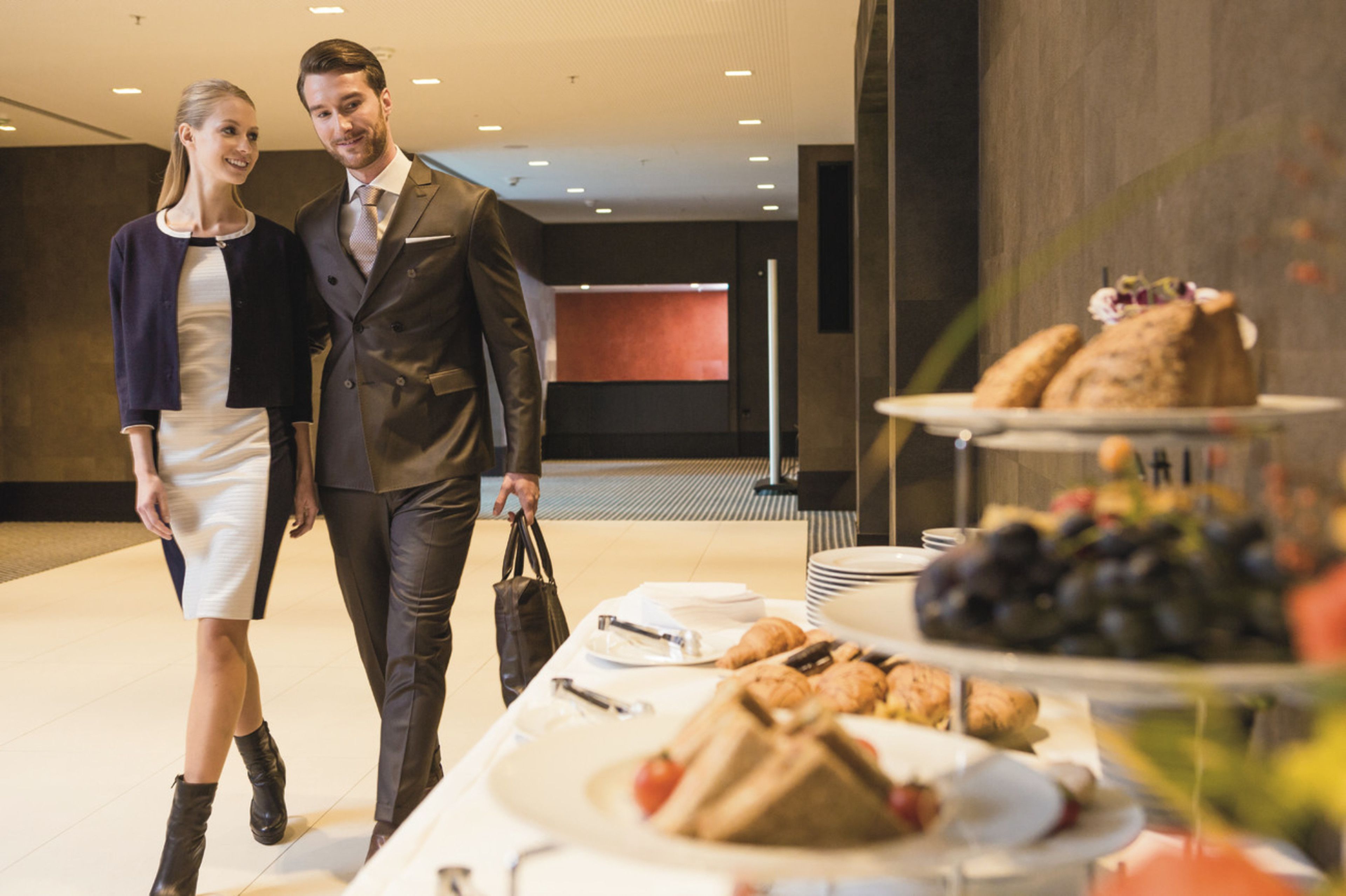 Un homme et une femme se promènent dans le hall d’un hôtel moderne, où se trouve une table de buffet proposant des sandwichs, des fruits et des pâtisseries.