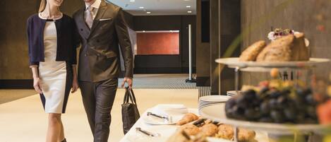 Un hombre y una mujer caminando en un moderno vestíbulo de hotel con una mesa de buffet que ofrece sándwiches, frutas y pasteles.