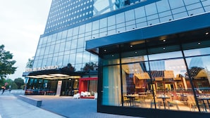 Un hotel moderno con fachada de vidrio, zona de estar al aire libre y un restaurante visible en su interior.