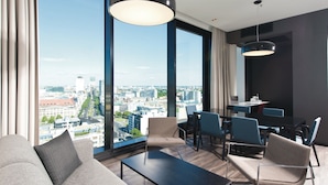 Habitación de hotel moderna con un ventanal amplio que ofrece vistas a la ciudad, un sofá, una mesa de comedor con sillas y una lámpara colgante.