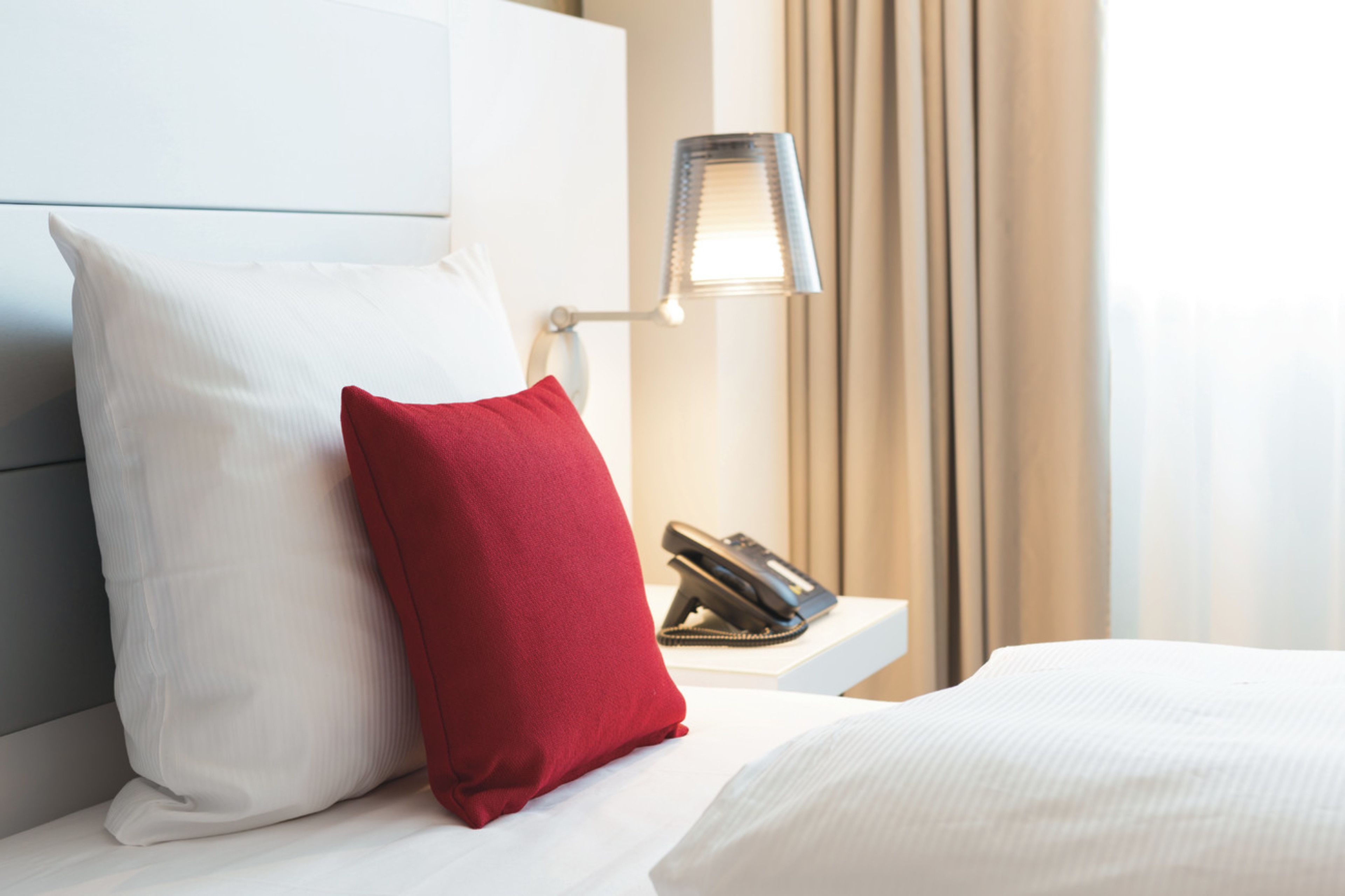 A hotel room with a bed, a red pillow, a white pillow, a nightstand with a telephone, and a lamp.