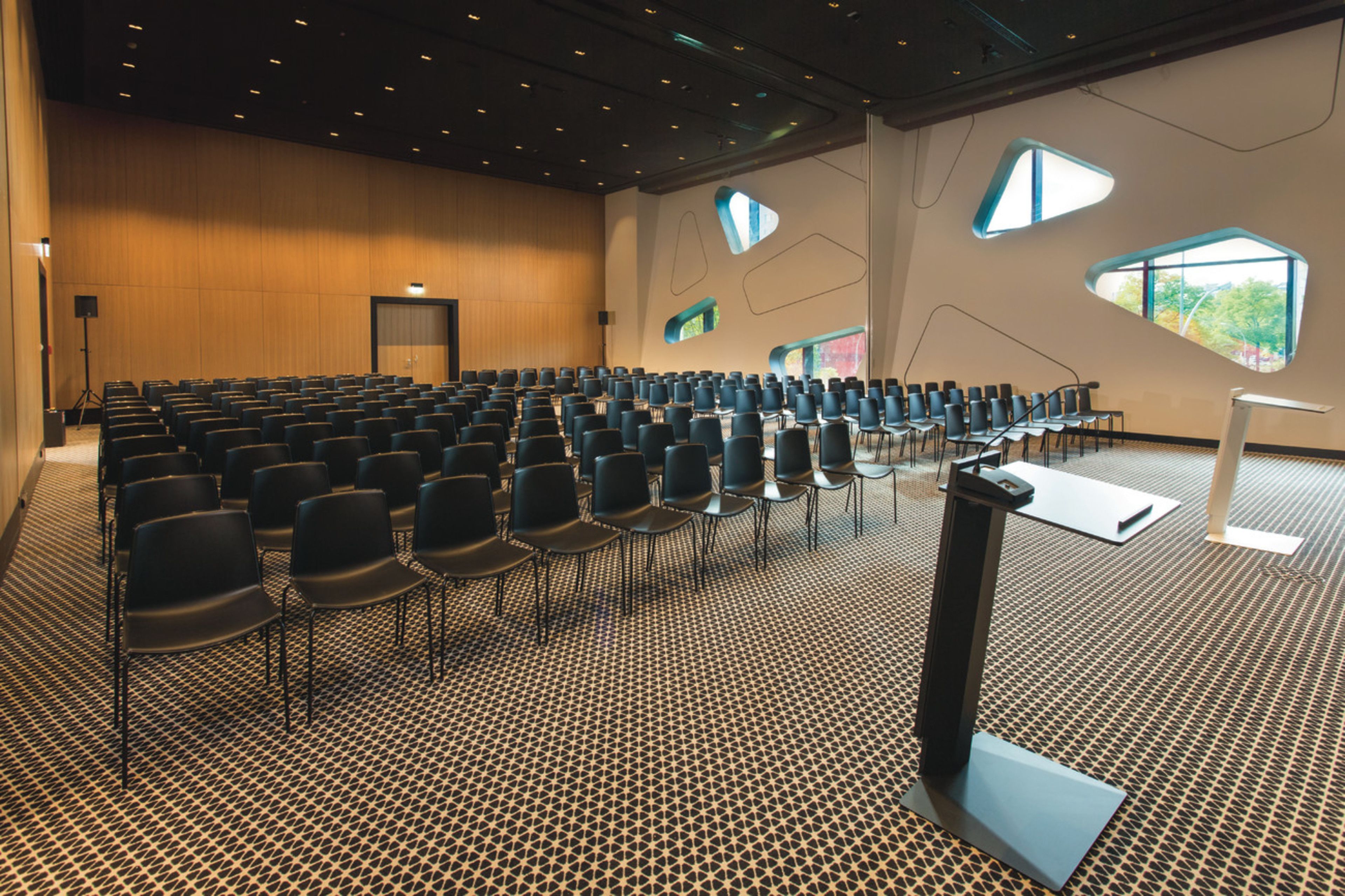A conference room with rows of chairs, a podium, and large windows with geometric shapes.