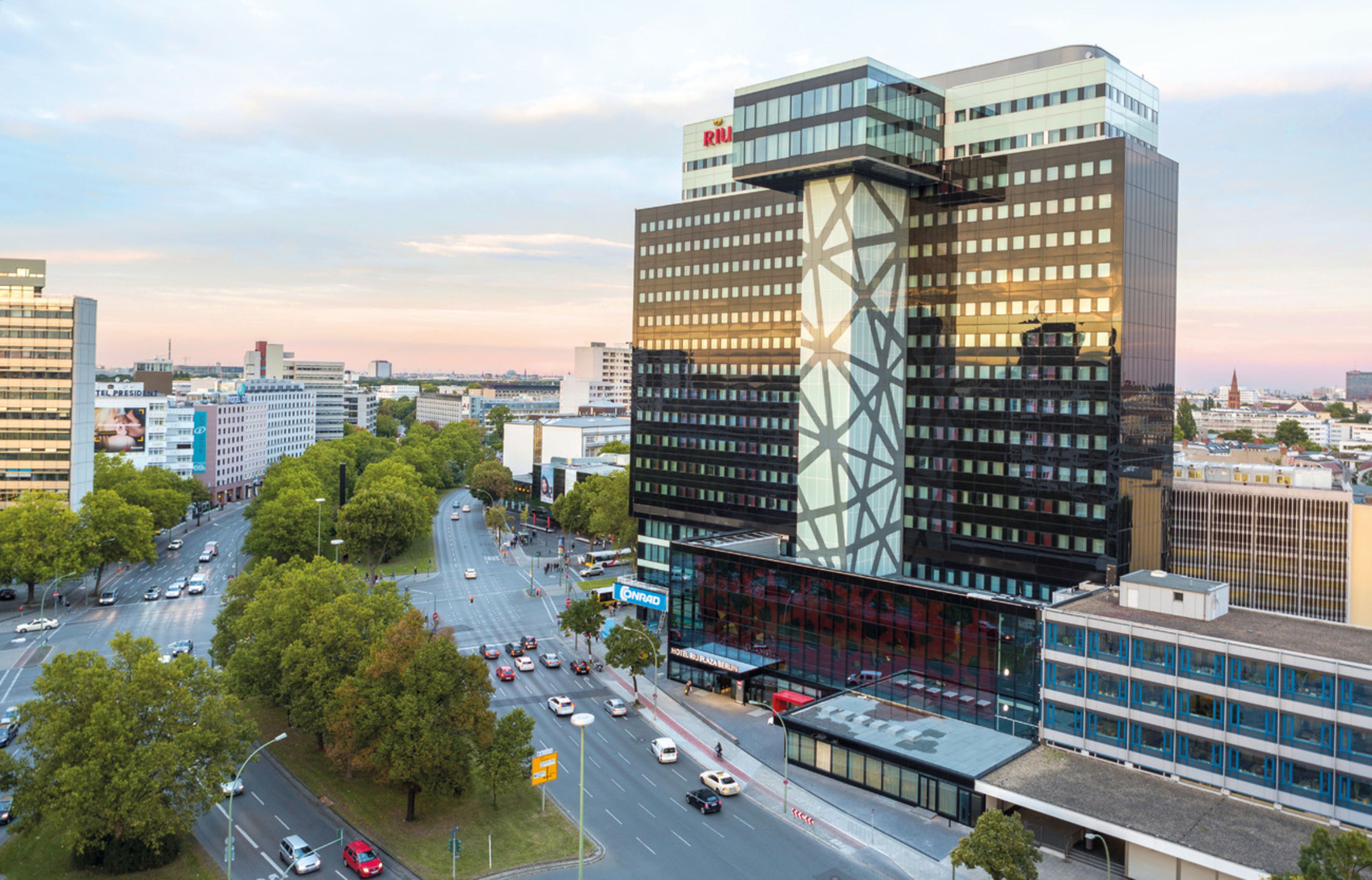 Un bâtiment urbain moderne à la façade vitrée, entouré d’une rue animée où circulent des voitures et où l’on trouve des arbres.