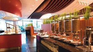 Un bufé moderno de hotel con un mostrador curvo, distintas estaciones de comida y plantas decorativas.