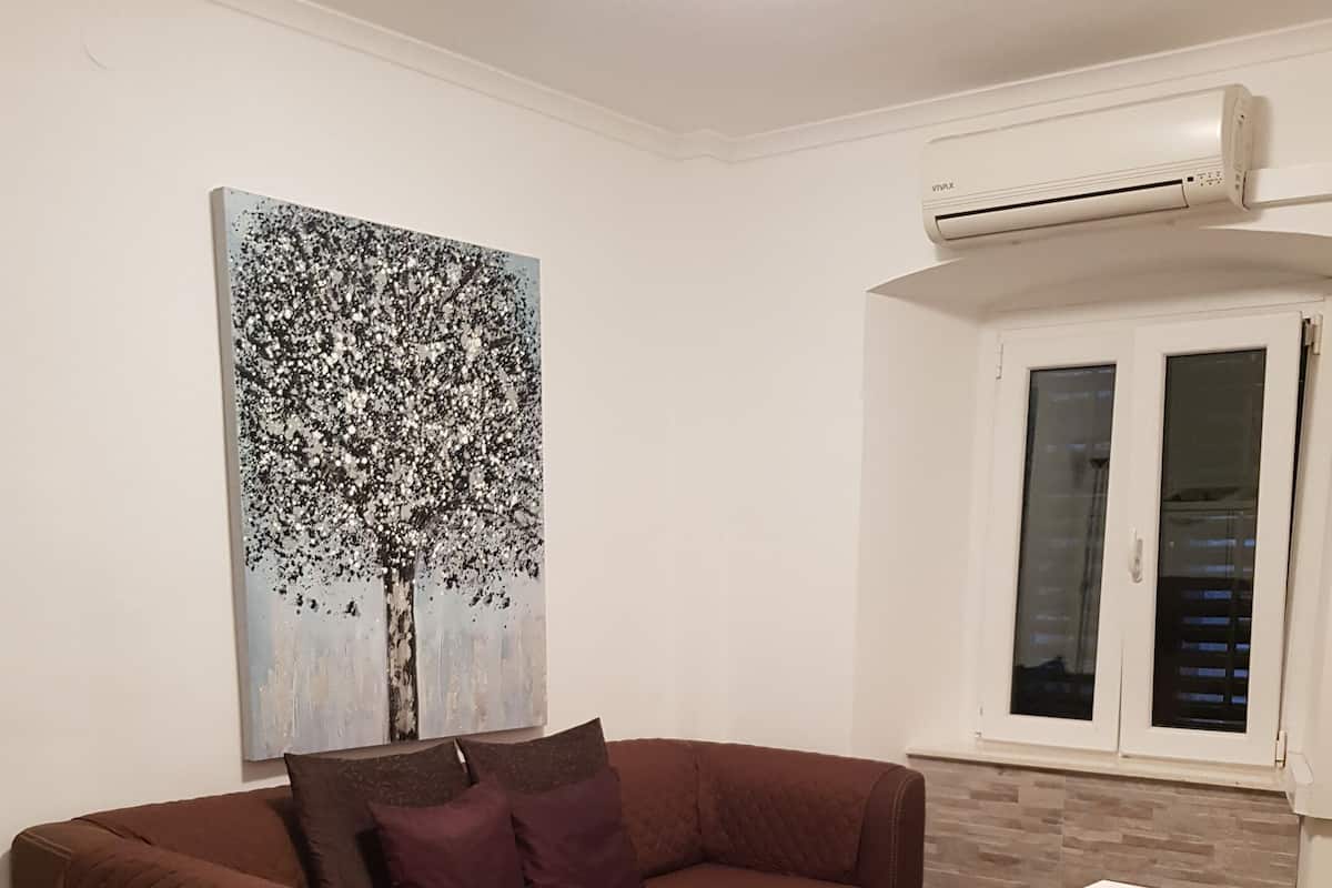 A living room with a brown sofa, a painting of a tree, a white table, and a window with blinds.