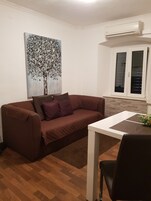 Uma sala de estar com um sofá marrom, uma pintura de uma árvore, uma mesa branca e uma janela com persianas.